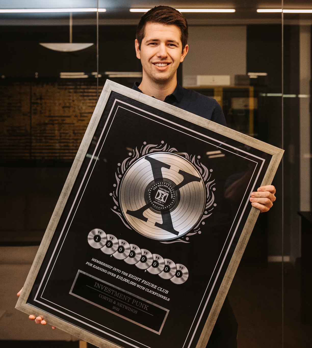 Gregor Heritsch hält im Büro den gerahmten ClickFunnels Two Comma Club X Award, eine schwarze Platte mit silbernem X