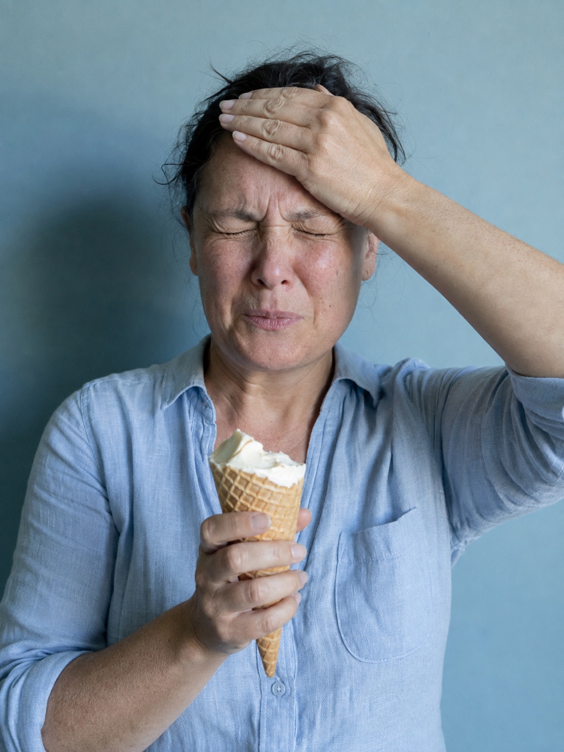 A woman wincing with her hand pressed to her forehead, holding an ice cream cone